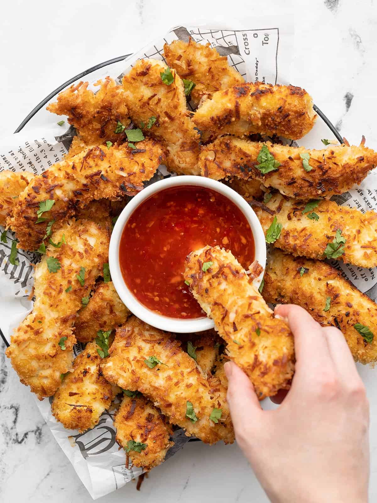 Overhead view of a plate full of coconut chicken strips with one being dipped into a bowl of sweet chili sauce