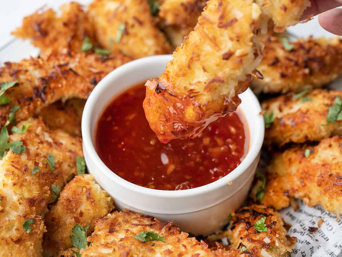 Close up of a coconut chicken strip being dipped into sweet chili sauce