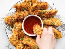 A plate of coconut chicken strips with a bowl of sweet chili sauce in the center, a hand dipping one chicken strip into the sauce