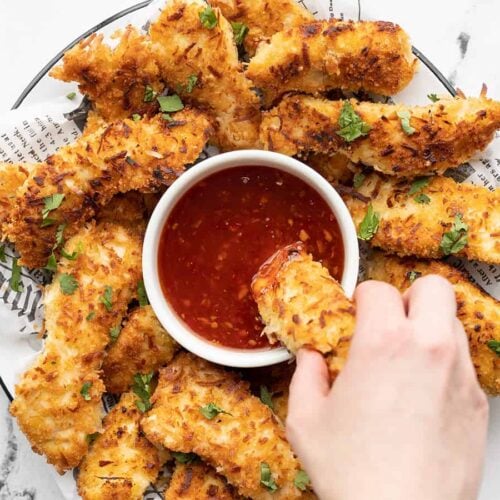 A plate of coconut chicken strips with a bowl of sweet chili sauce in the center, a hand dipping one chicken strip into the sauce