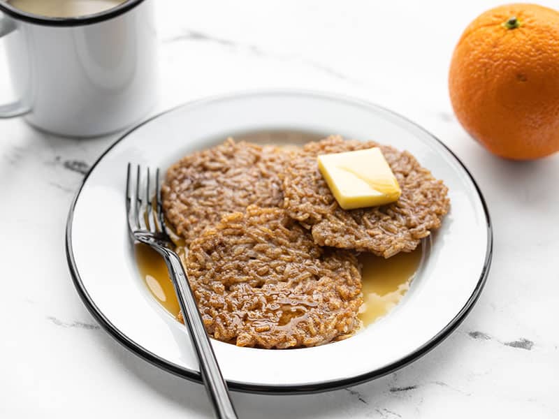 Front view of rice pancakes on a plate with butter and maple syrup, an orange and cup of coffee in the back