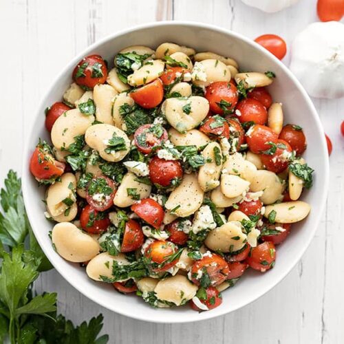 Overhead view of a bowl full of Mediterranean White Bean Salad with parsley, tomatoes, and garlic on the side