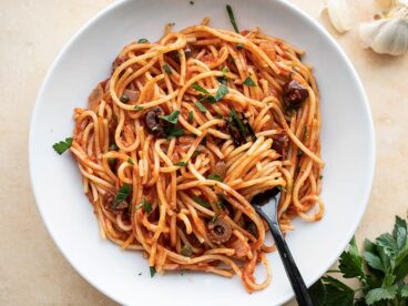 A bowl of Pasta Puttanesca with a fork twirled in the center. Garlic and parsley on the side.