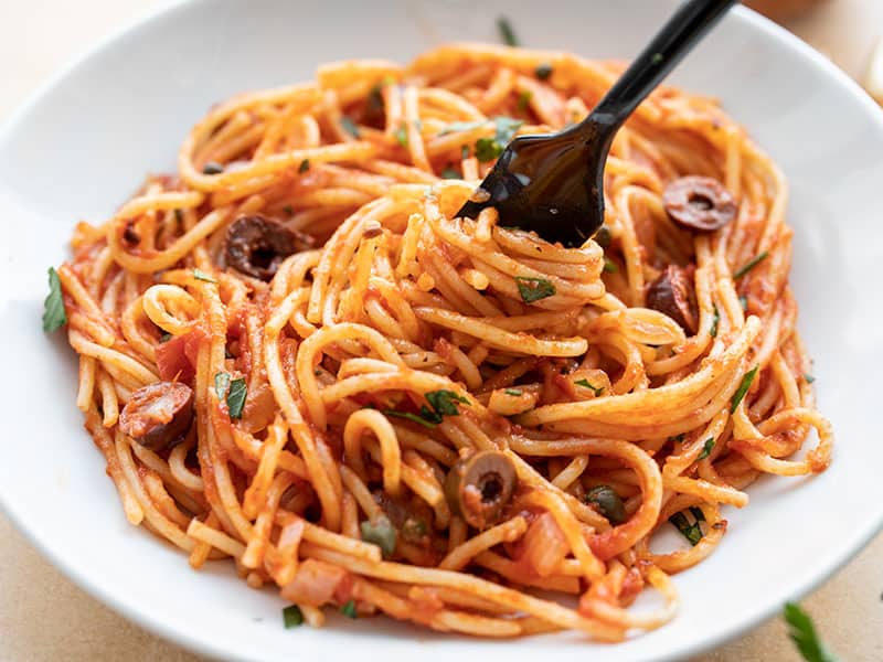 Front view of a bowl of Pasta Puttanesca with a fork twirling the pasta.