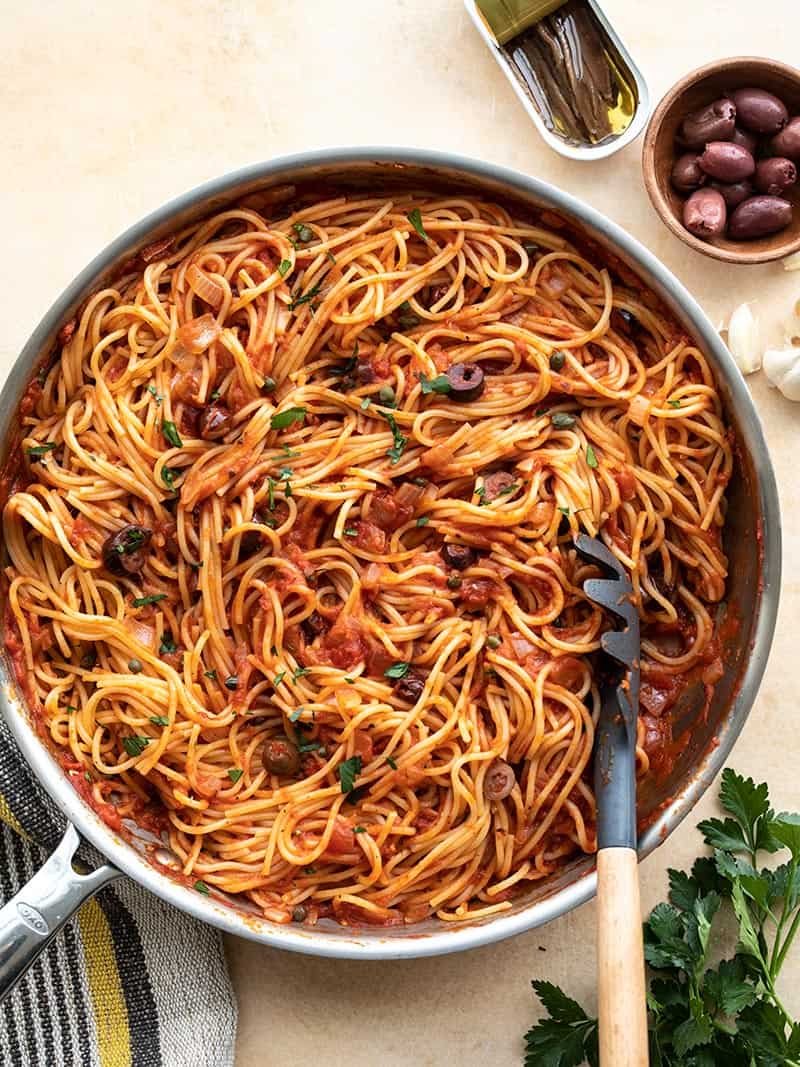 A large skillet full of pasta puttanesca, with a pasta fork in the side, anchovies and olives next to the skillet.