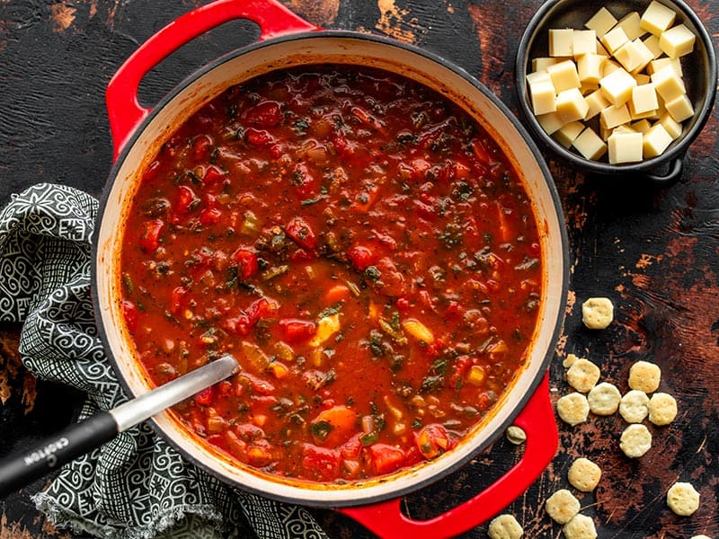 A red soup pot full of Swamp Soup with a bowl of Swiss cheese cubes on the side