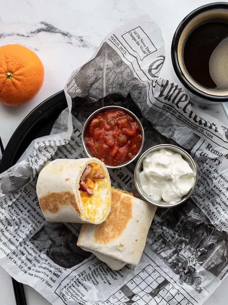 A freezer breakfast burrito cut open on a plate with little cups of salsa and sour cream, a cup of coffee and a tangerine on the side.