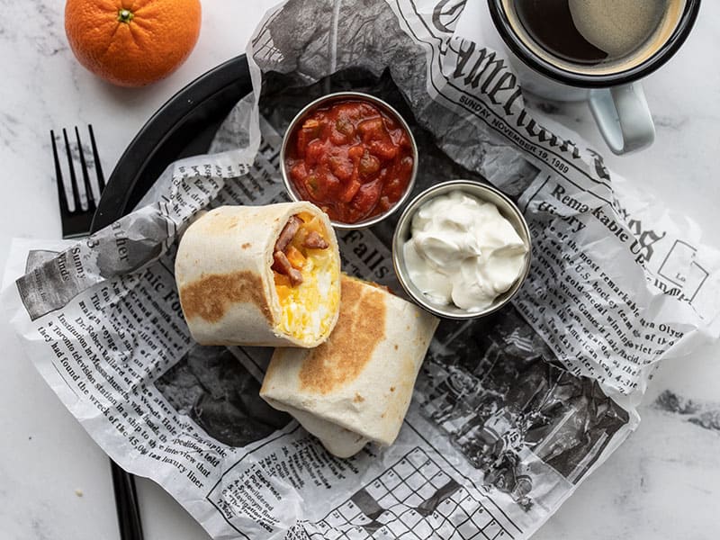 A reheated breakfast burrito on a plate with a newsprint liner, two small dishes of salsa and sour cream, a tangerine and cup of coffee on the side