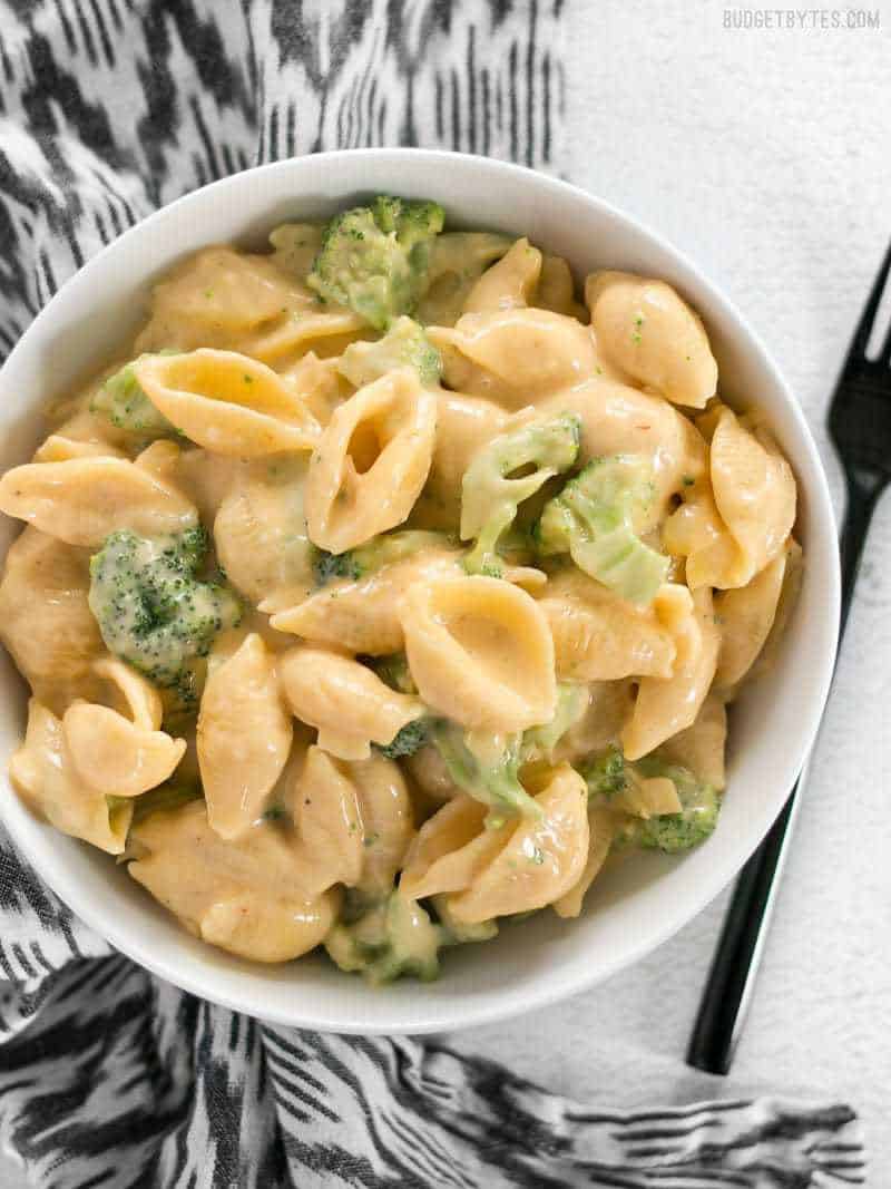 top view of a bowl of broccoli shells n' cheese sitting on a black and white patterned napkin with a fork on the side 