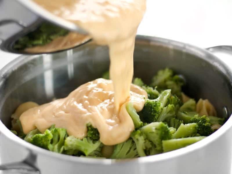 Pouring cheese sauce over cooked noodles and broccoli in pot 