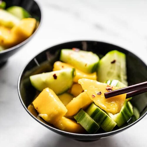 Close up of a bowl of cucumber mango salad with chopsticks picking up a piece of mango