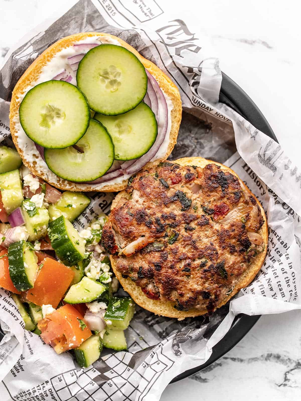 Overhead view of a mediterranean turkey burger, top bun off, on a paper lined plate next to cucumber salad