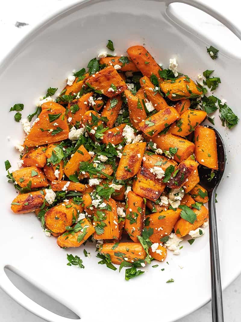 A white serving bowl full of Roasted Carrot and Feta Salad, a black spoon in the side.