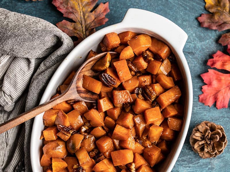 Maple Roasted Sweet Potatoes in a casserole dish with a wooden spoon on a blue background with leaves and pine cones on the side.