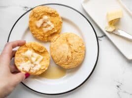 A hand picking up half of a sweet potato biscuit, smeared with butter and drizzled with maple syrup