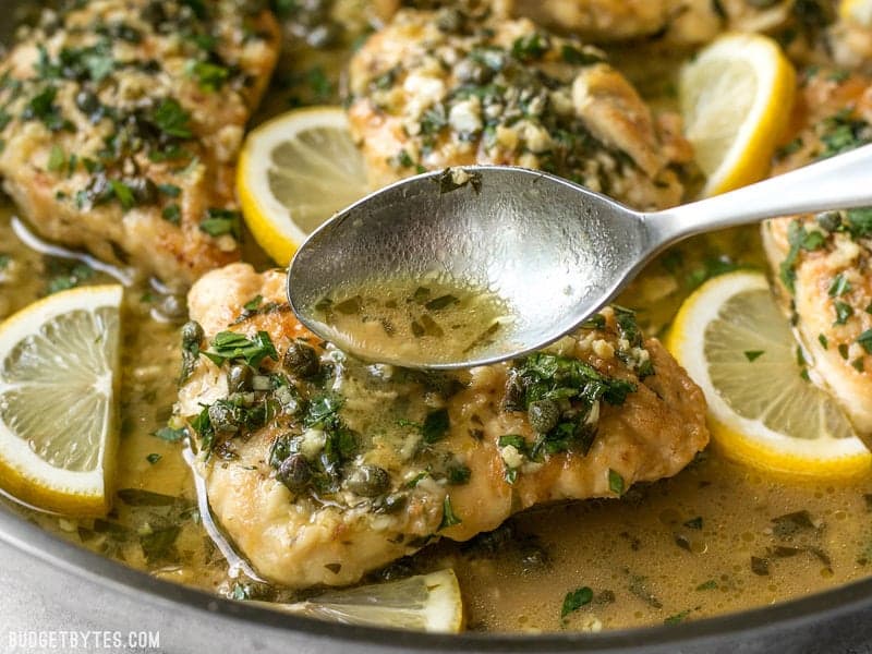 Lemon garlic pan sauce being drizzled over a piece of Chicken Piccata with capers and parsley.