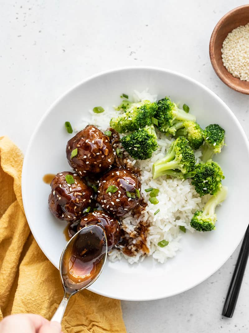 Teriyaki meatball bowls with teriyaki sauce being drizzled on top with a spoon, a bowl of sesame seeds on the side.