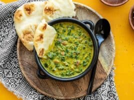 A black bowl with Indian Creamed Spinach on a wooden plate with a piece of naan in the side of the bowl