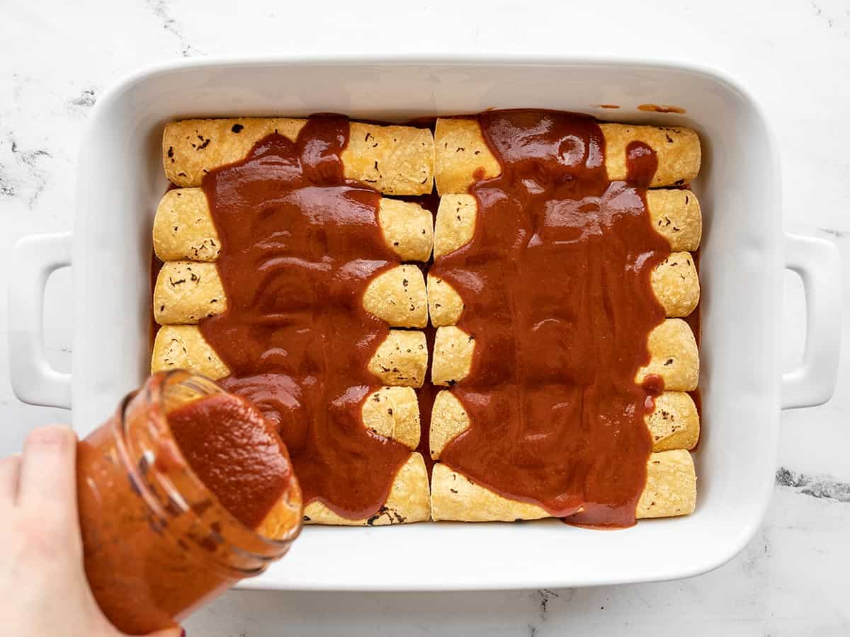 enchilada sauce being poured from a glass jar over a casserole dish full of enchiladas.