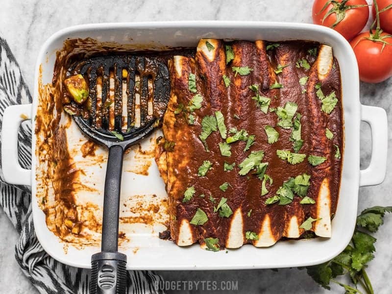 Casserole dish Black Bean and Avocado Enchiladas, some have been served, spatula resting in their place