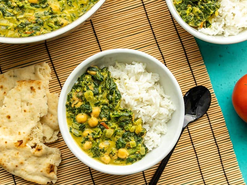 Two bowls of chana saag with rice, next to the skillet and a piece of torn naan