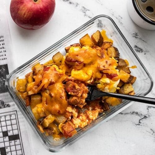 One Country Breakfast Bowl in a glass container being eaten with a black fork next to a newspaper, coffee mug, and apple