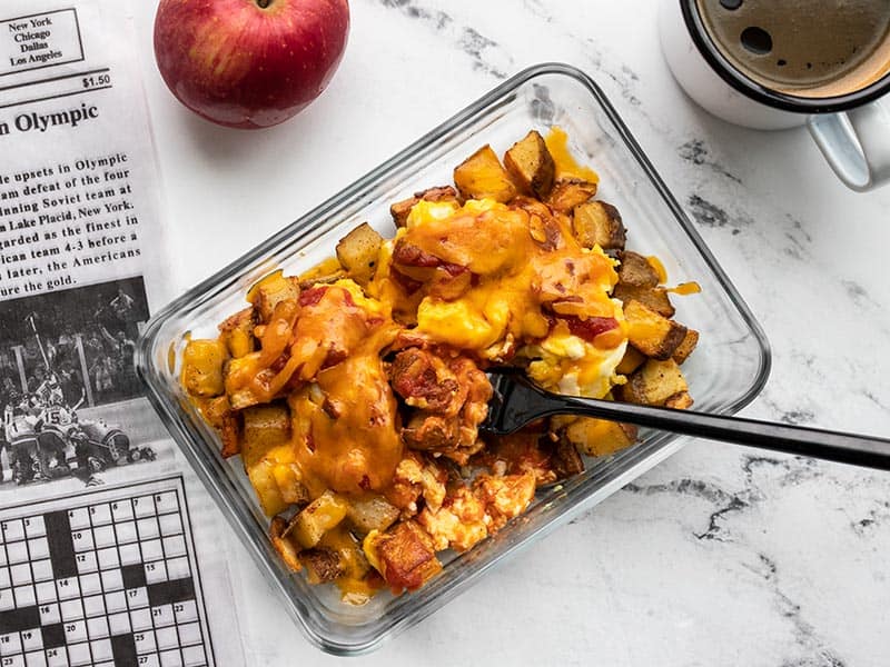 One Country Breakfast Bowl in a glass container being eaten with a black fork next to a newspaper, coffee mug, and apple