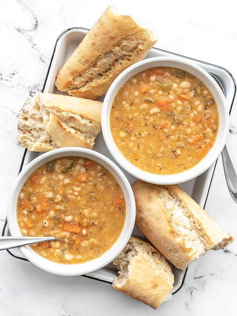 Overhead view of two bowls of slow cooker white bean soup on a white tray with torn pieces of baguette on the sides