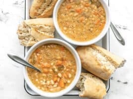 Two bowls of white bean soup on a white tray with torn baguette pieces on the side