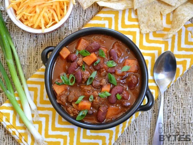 Top view of a bowl of Chorizo Sweet Potato Chili with a spoon and bowl of shredded cheese on the side 