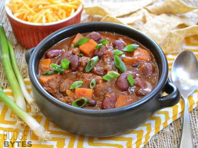 Side view of a bowl of Chorizo Sweet Potato Chili with a spoon and a bowl of shredded cheese on the side 