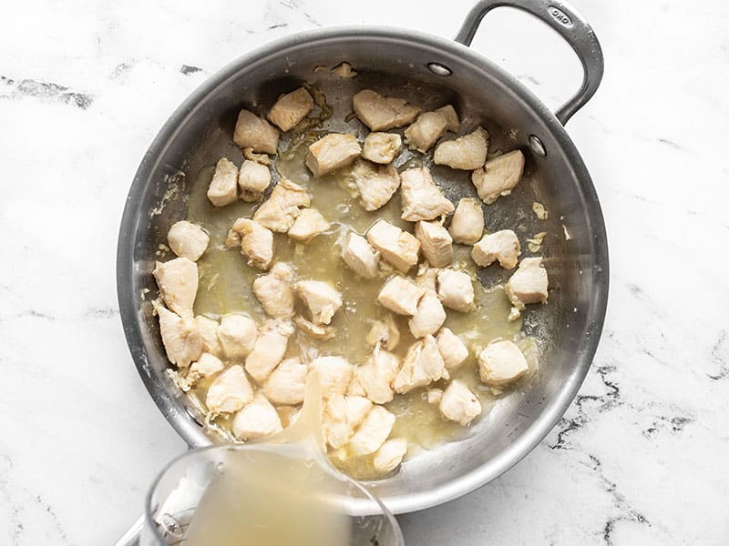 Broth being poured into a skillet with cooked chicken pieces.