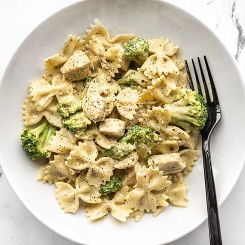 A bowl full of creamy pesto pasta with chicken and broccoli, a black fork on the side