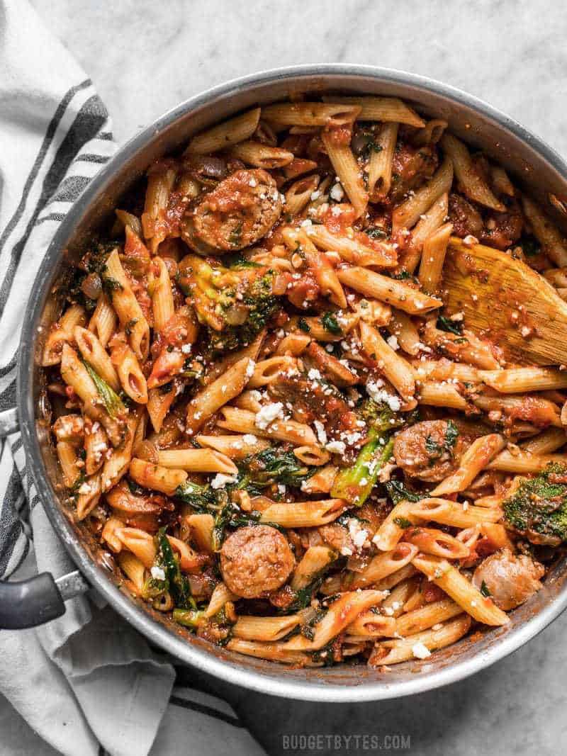 A large pot full of Penne Pasta with Sausage and Greens with feta sprinkled over top and a wood spoon