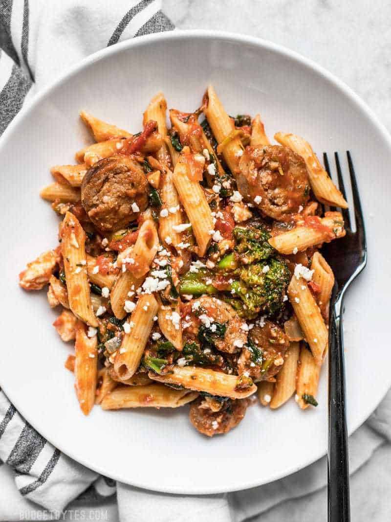 A white plate full of Penne Pasta with Sausage and Greens, and a black fork. 