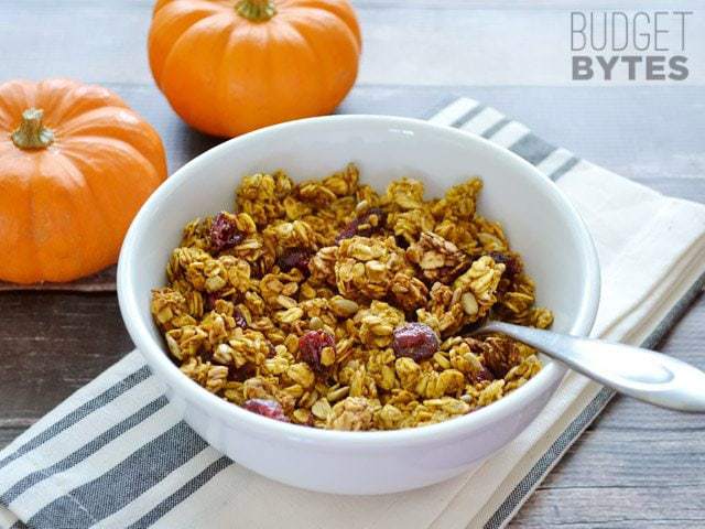 Bowl of Pumpkin Spice Granola withe a spoon, two small pumpkins and a gray and white napkin for decor 