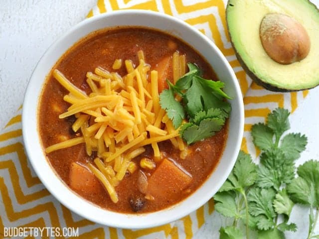 Top view of a bowl of Sweet Potato Tortilla Soup, shredded cheese and cilantro on top. Yellow chevron napkin, half an avocado and cilantro on the side for staging. 