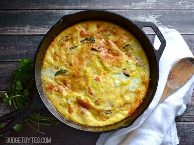 Top view of a baked Zucchini and Corn Frittata, wooden spoon, white napkin and cilantro on the side 