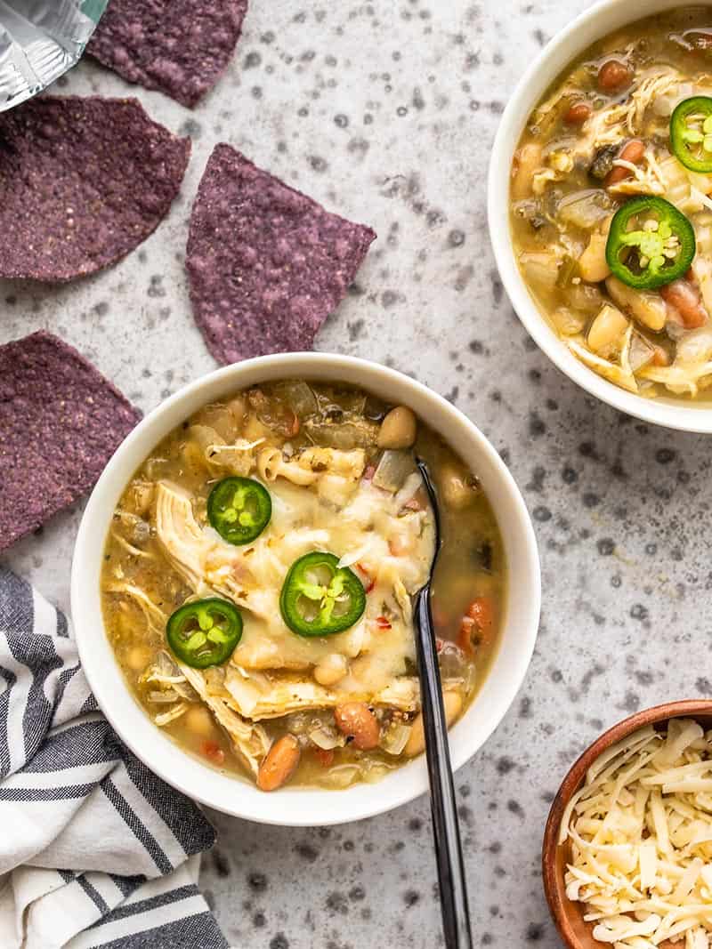 Two bowls of slow cooker white chicken chili topped with cheese and blue tortilla chips on the side