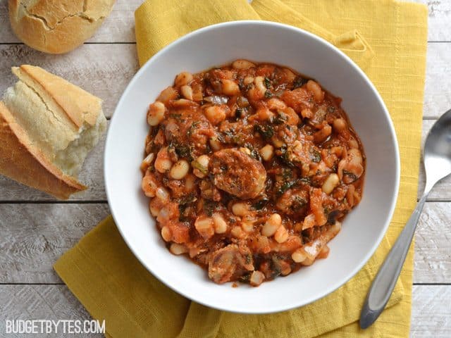 Top view of a bowl of White Beans with Tomato and Sausage sitting on a yellow napkin, fork and slices of bread on the side 