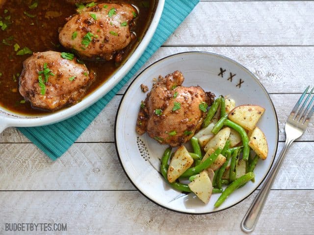 Dish of Balsamic Chicken Thighs with one thigh dished onto a plate with a side of potatoes and green beans 