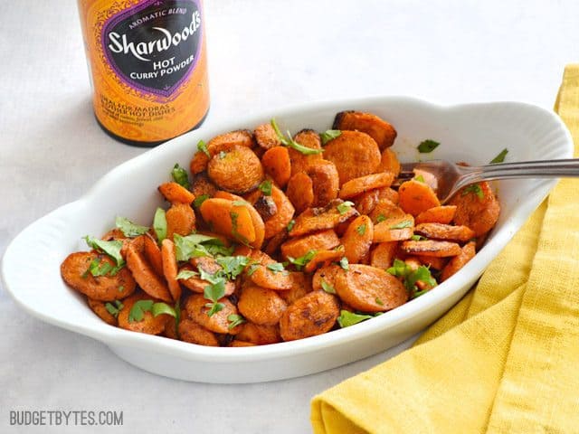 Dish of cooked Curry Roasted Carrots, yellow napkin and bottle of curry powder on the side 