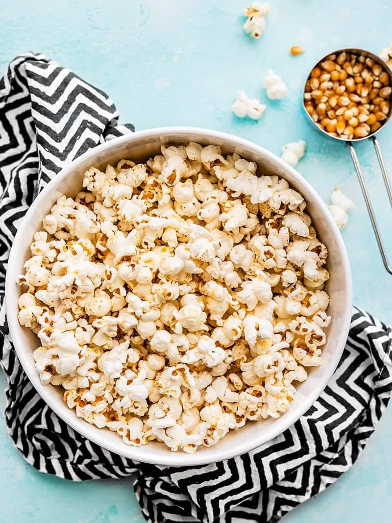 A big bowl of stovetop popcorn on a zig zag napkin with a measuring cup of unpopped kernels on the side