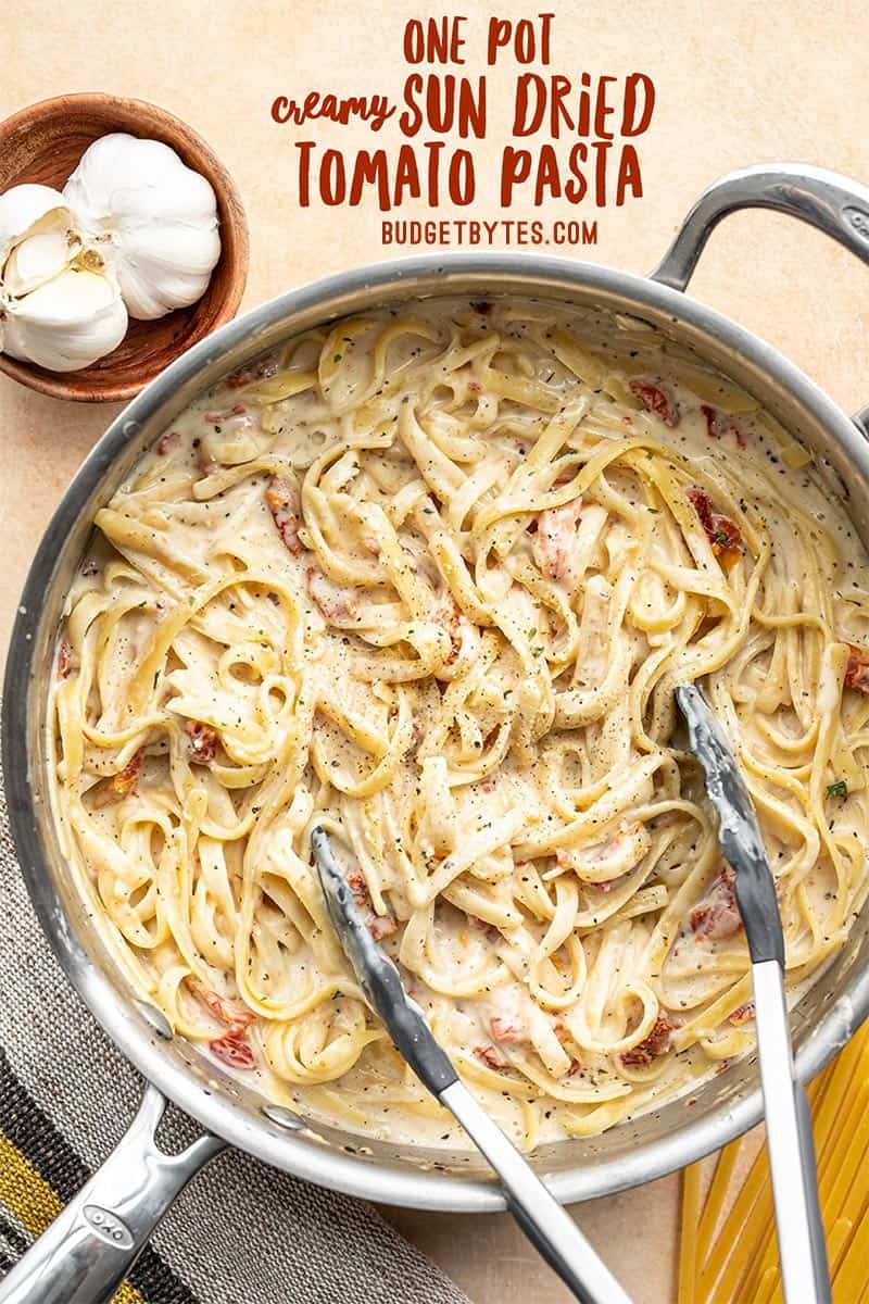 Overhead view of creamy sun dried tomato pasta in the skillet with tongs. Title text in the top corner.