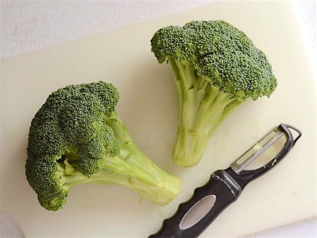 Two crowns of broccoli on a cutting board with a vegetable peeler