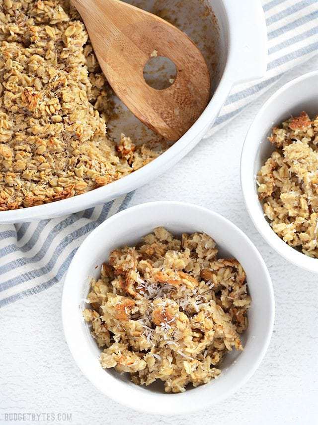 Banana Coconut Baked Oatmeal served into two bowls, casserole dish with a wooden spoon on the side