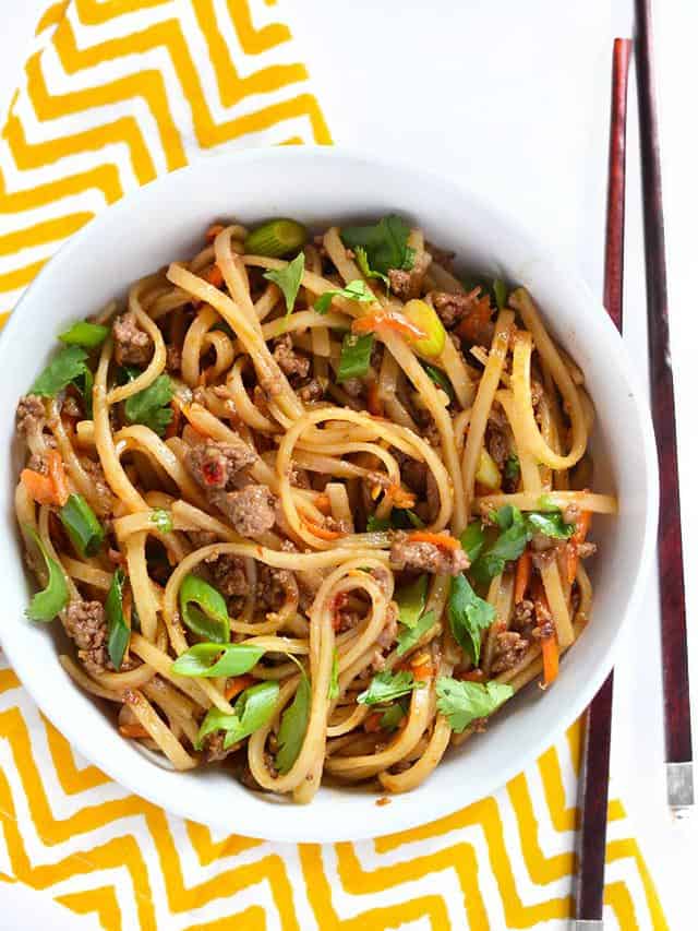 Overhead view of a big bowl of beef stir fry noodles on a yellow and white napkin, chopsticks on the side