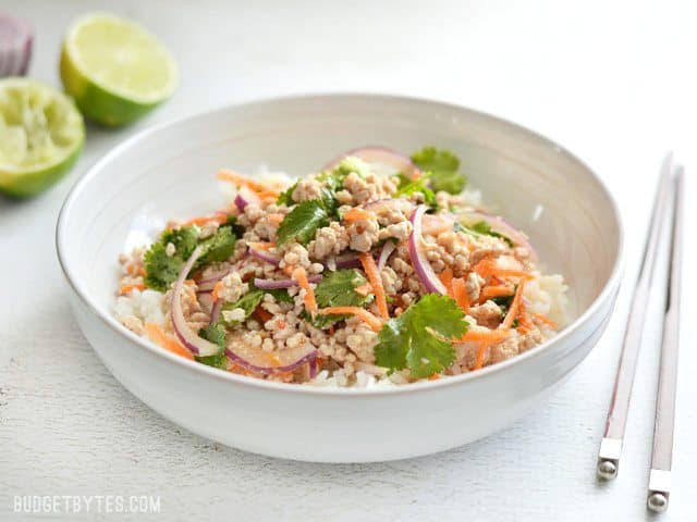 Side view of a bowl full of nam sod (Thai Pork Salad), with limes in the background and chopsticks on the side