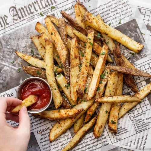 a hand dipping a fry into a cup of ketchup next to a pile of fries