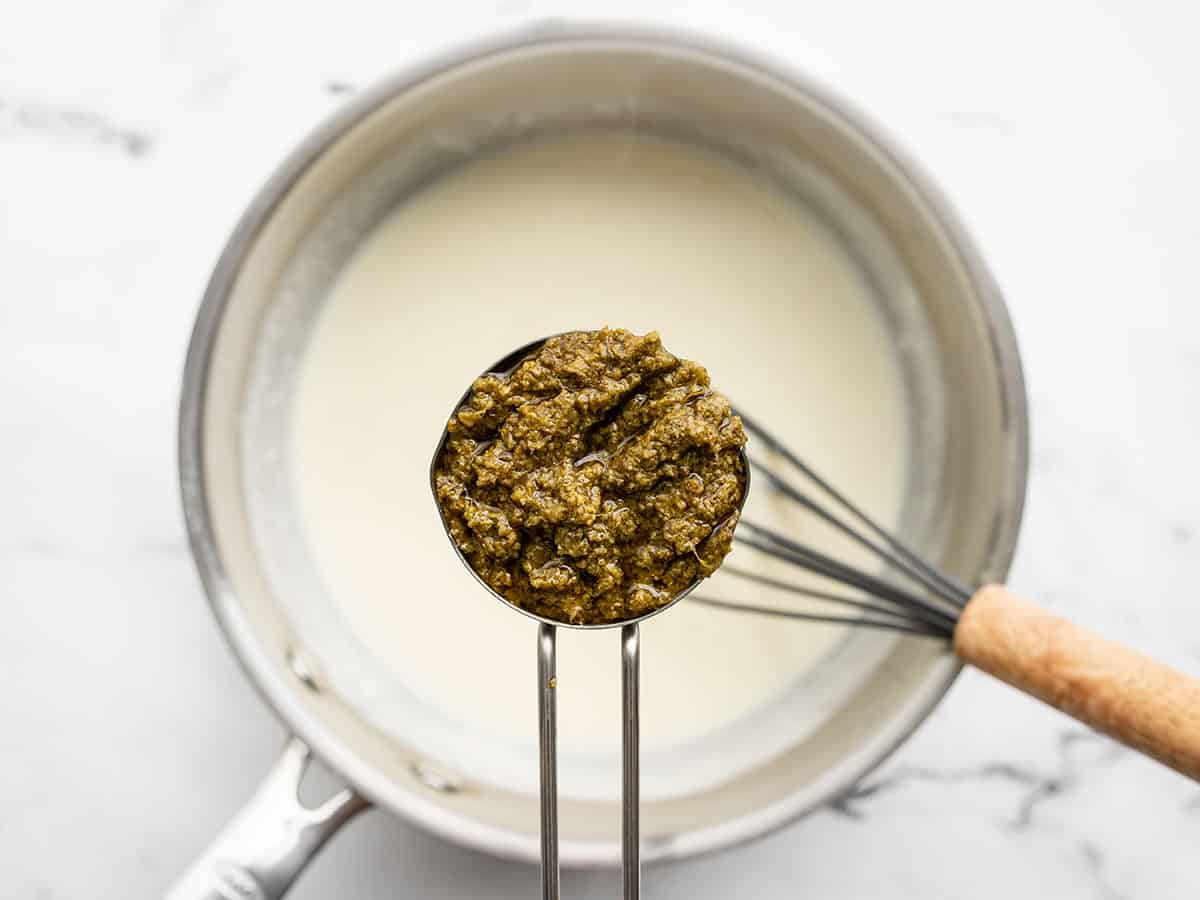 A measuring cup full of basil pesto held over the sauce pot